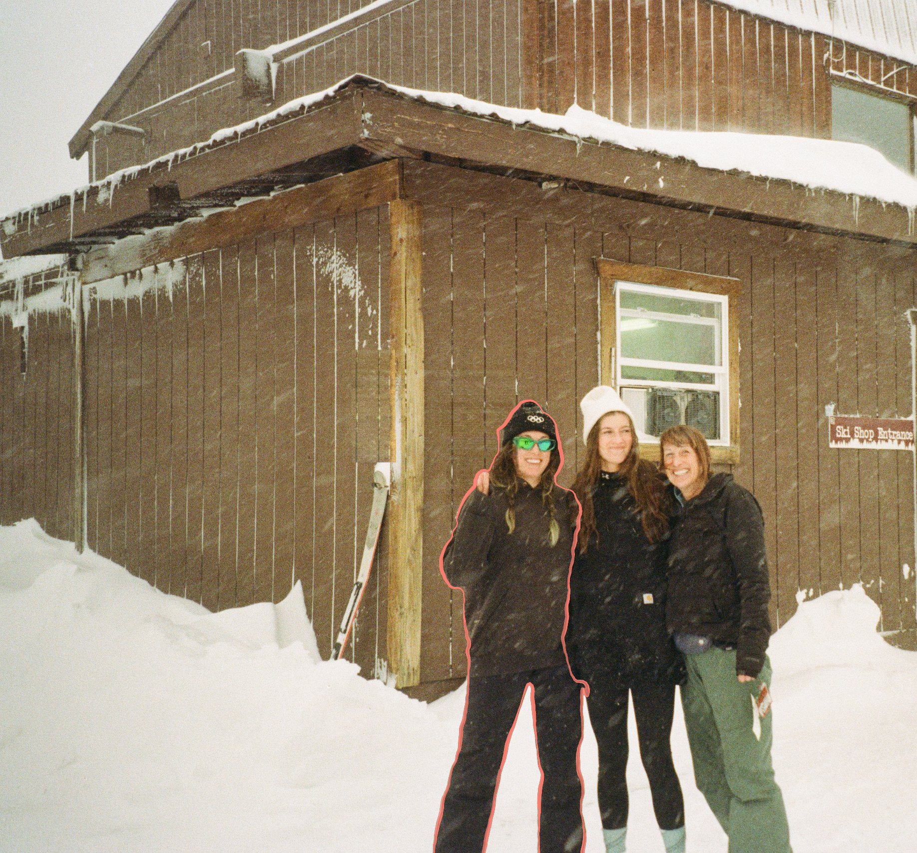 Three people smiling in the snow outside a wooden ski lodge; the person on the left is traced with a thin hand-drawn salmon-pink line