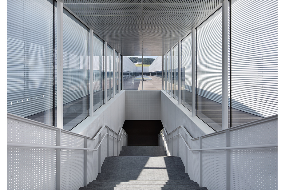 Stairwell between white perforated railings, with a glazed corridor and deck beyond