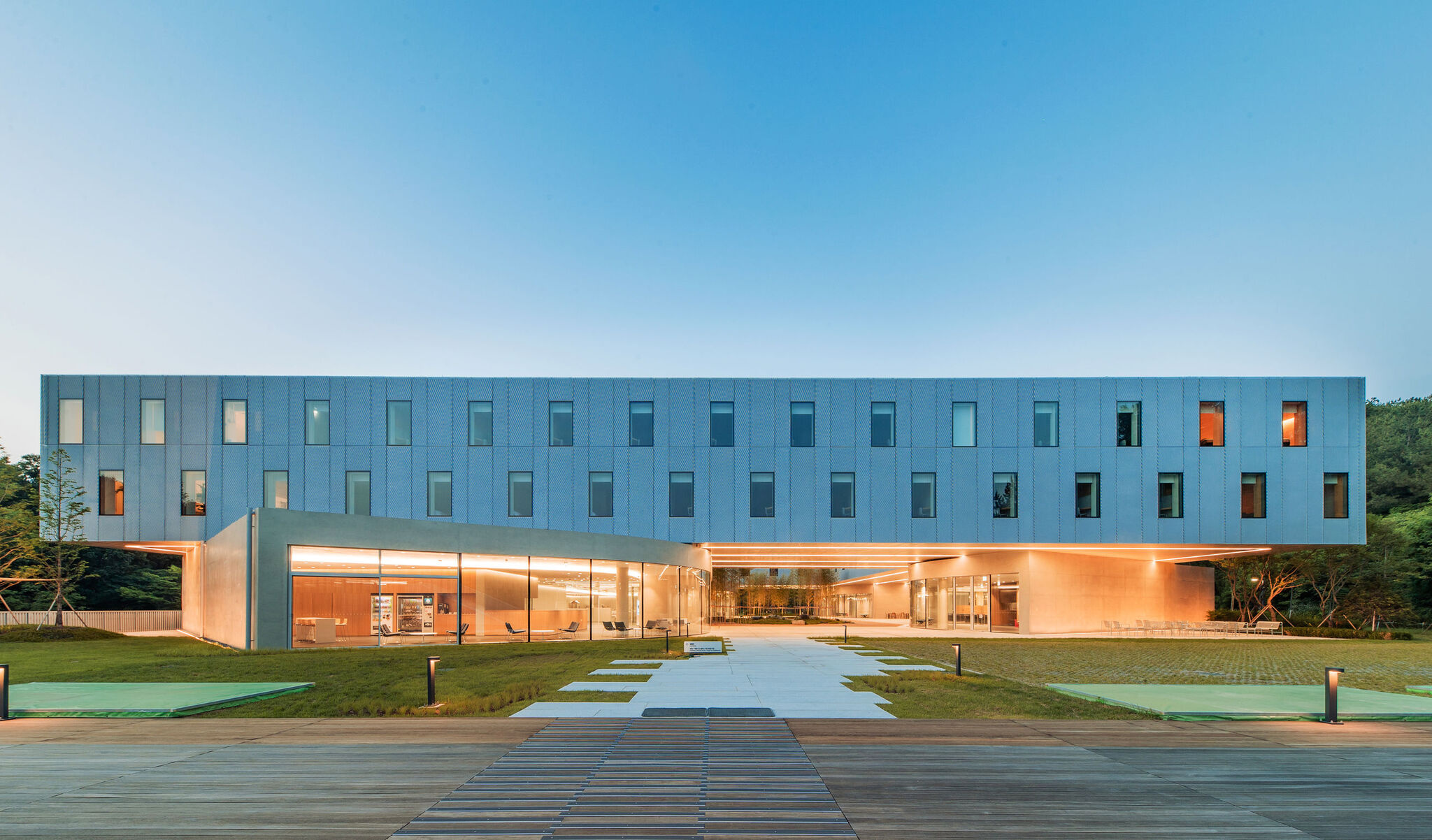 The Hyundai Cheonan Global Learning Center at dusk: a silver mesh volume floating above a glowing glass ground floor, with a paved path and lawn in front