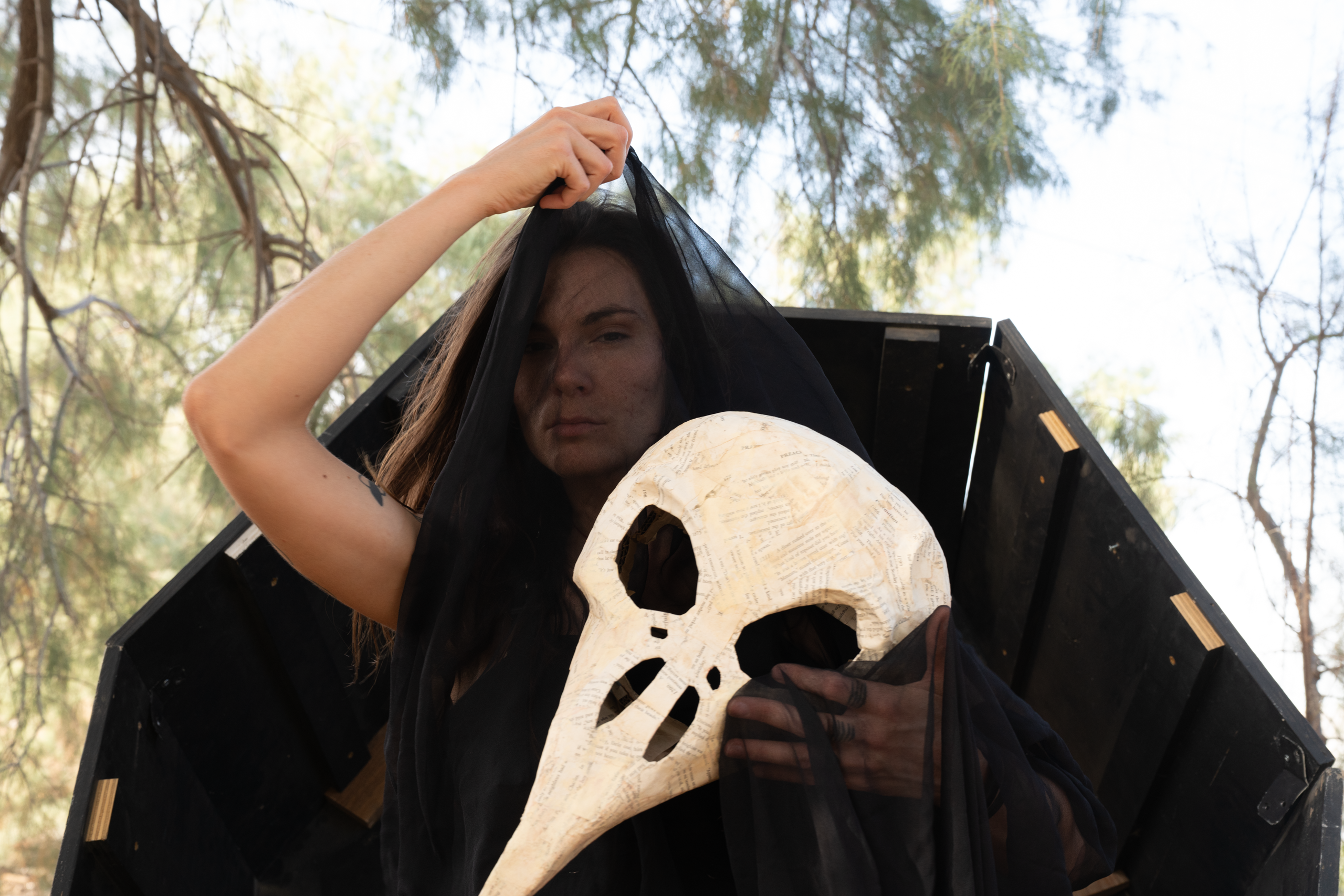 Person in a sheer black veil holding a paper-mâché bird-skull mask in front of a black wooden structure